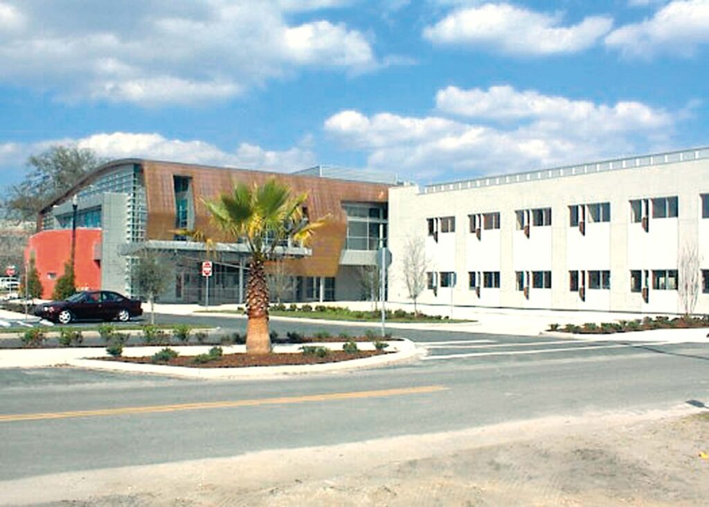 A modern medical office with a mix of red and white facades features large windows and a curved roof. A single palm tree stands gracefully in front, alongside a parked car. The partly cloudy sky adds charm to this contemporary space.