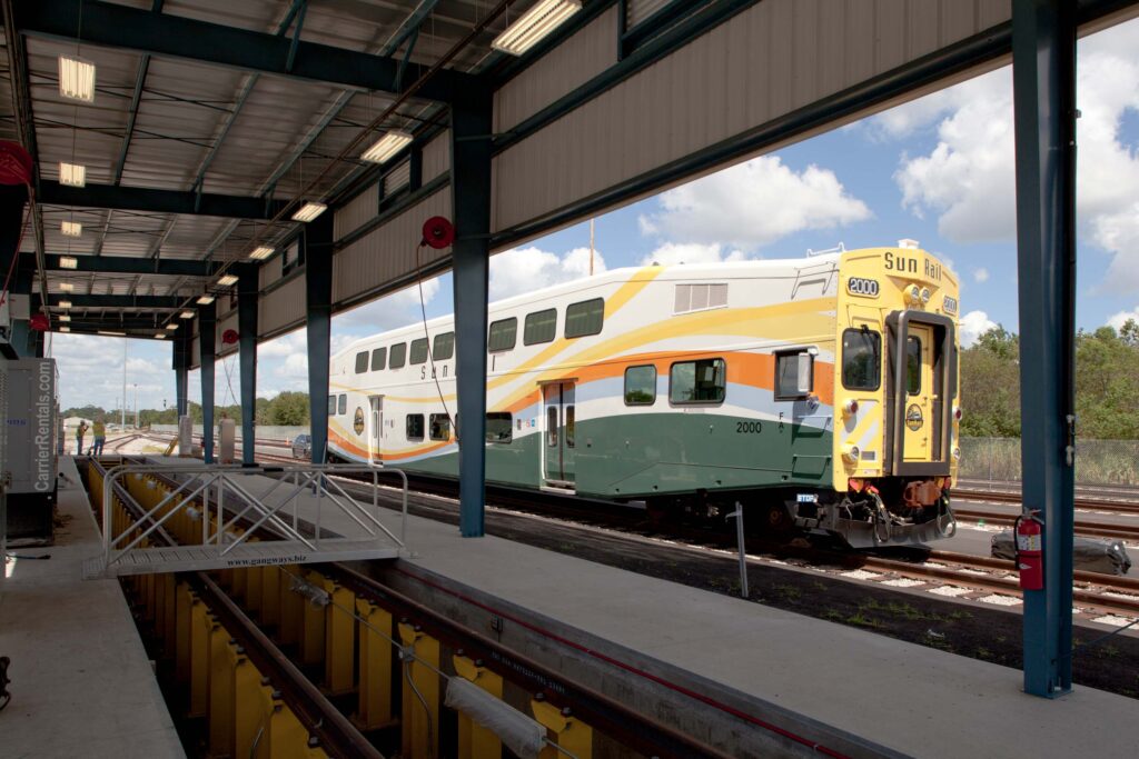 A commuter train at the Service and Inspection Facilities is positioned on elevated tracks under a covered structure, with control panels and safety barriers visible, part of the efficient operations in Sunrail Phase 1.