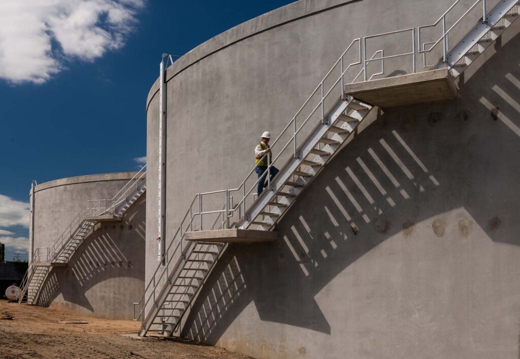 A person in a hard hat and safety vest climbs stairs on a large concrete industrial tank in Curtis Bay, implementing stormwater reclaim upgrades under a clear blue sky.