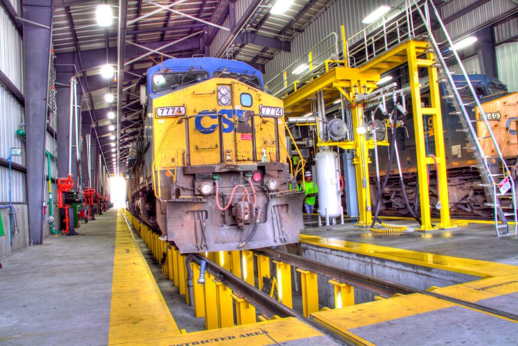 A yellow and blue CSX locomotive numbered 7774 sits inside the service center, with another train visible to the right. The area is equipped with large machinery and bright yellow platforms.