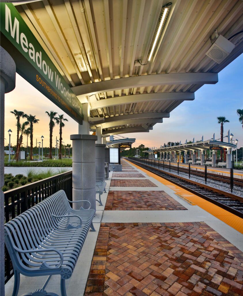 Empty train station platform with overhead shelters, benches, and illuminated signs at dusk. Palm trees line the background beneath a colorful evening sky, part of the Southern Expansion and Sunrail Phase 2, hinting at bustling days ahead for this Vehicle Service Facility.