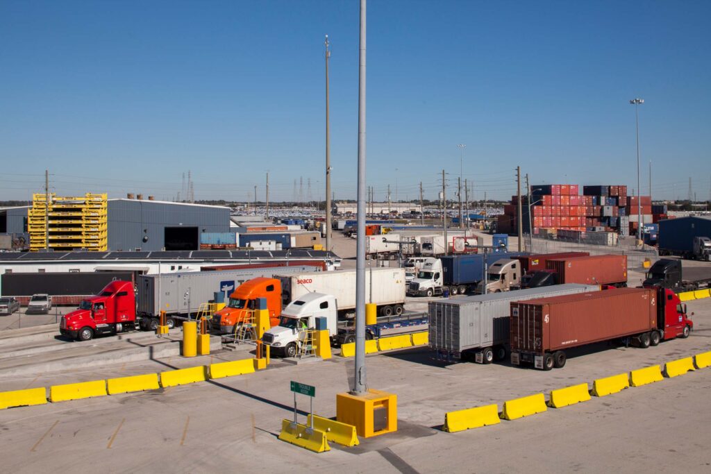 Freight trucks lined up at a revitalized shipping terminal feature Tote Maritime's branding, with stacked cargo containers and modern industrial buildings in the background. The scene is set against a clear sky in bright daylight.