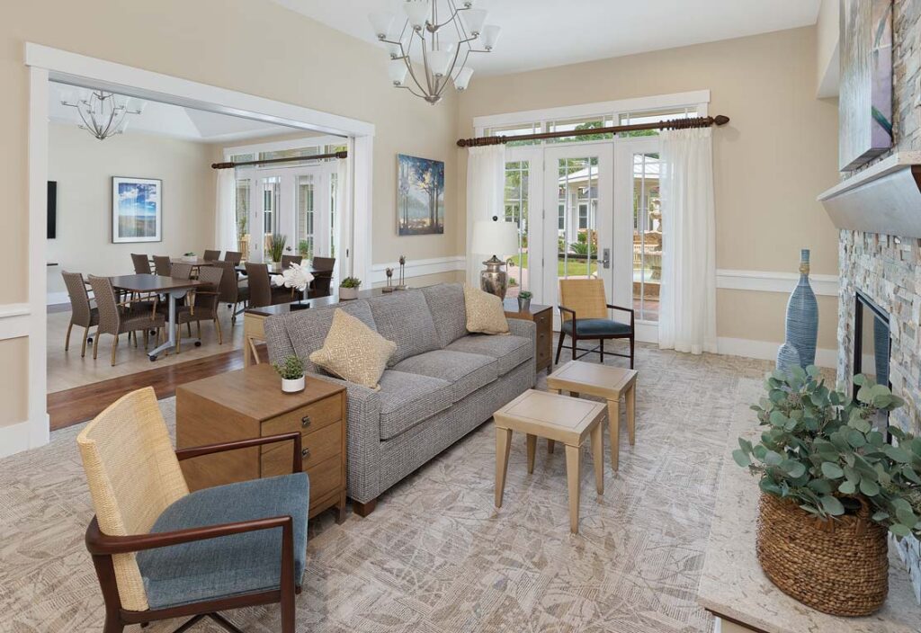 A spacious living room at Mount Pleasant Gardens, featuring a gray sofa, armchairs, and a stone fireplace. The dining room is visible in the background. Large windows bring in natural light, while neutral colors dominate the decor of this elegant assisted living facility.