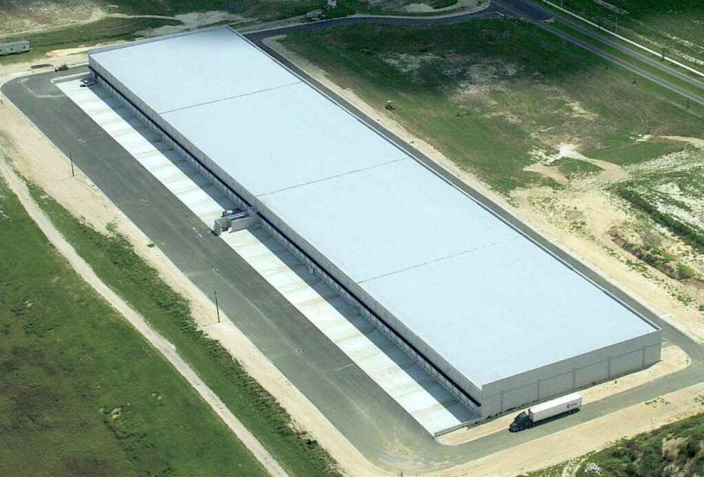 An aerial view of a warehouse framed by an expansive field. In the distance, a road meanders past. A single semi-truck approaches the loading dock in the foreground.