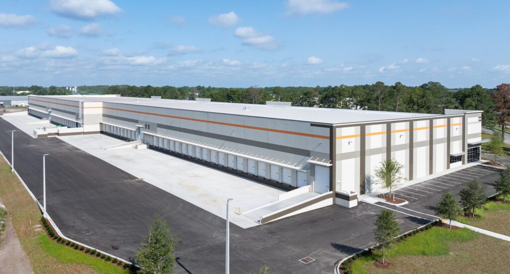 Aerial view of a large, modern warehouse in Cypress Plaza, featuring a flat roof and loading docks. It sits amidst an industrial park with a surrounding parking area, all set against a backdrop of trees under a partly cloudy sky.