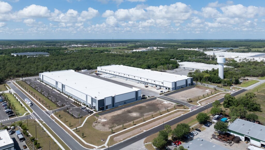 Aerial view of the Poinciana Logistics Center, showcasing Buildings 1 & 2 amidst expansive parking lots and greenery. In the distance, a water tower stands under a partly cloudy sky.