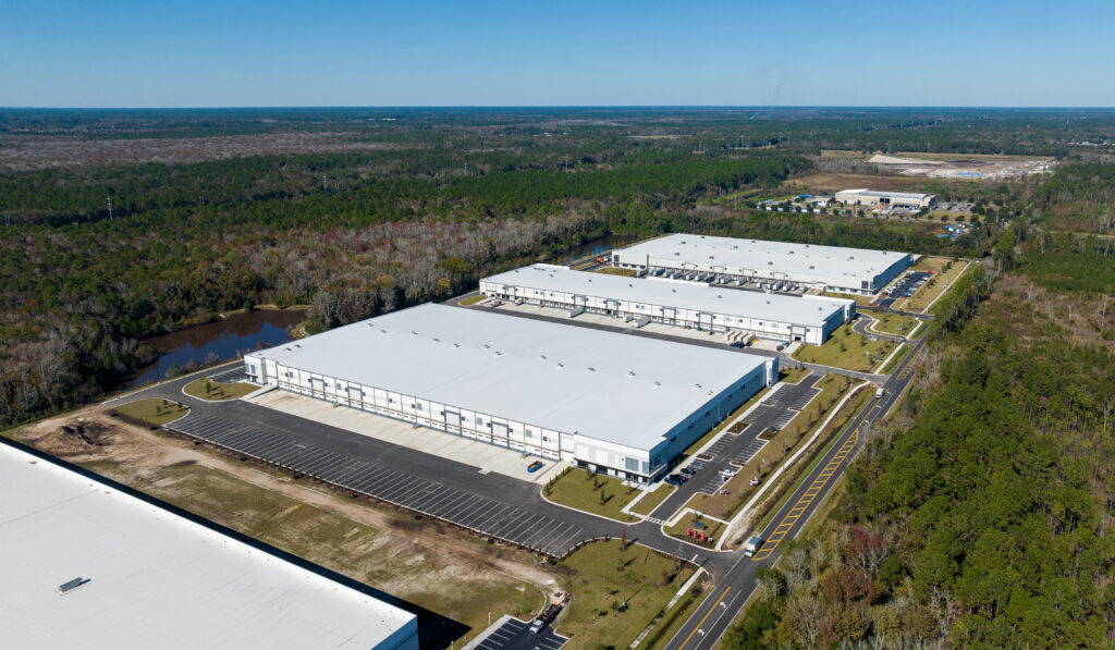 Aerial view of large white warehouse buildings surrounded by greenery and parking lots along Pattillo's Imeson Road under a clear blue sky.