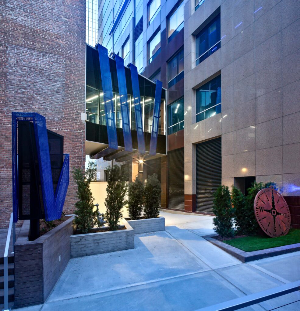 A bridge between modern buildings with reflective glass, featuring four large blue panels, leads to the contemporary elegance of Vystar Credit Union's West Bay branch.