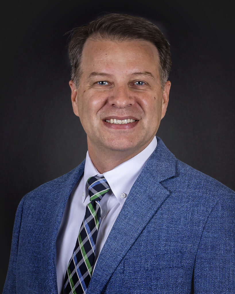 Jamison Chattin, dressed in a blue suit jacket, white shirt, and patterned tie, smiles at the camera against a dark background.