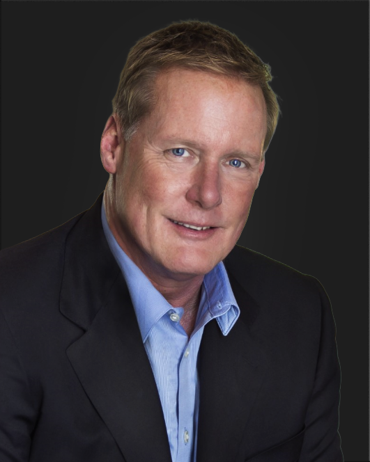 A man with short light brown hair in a dark suit jacket and light blue shirt poses against a plain dark background.