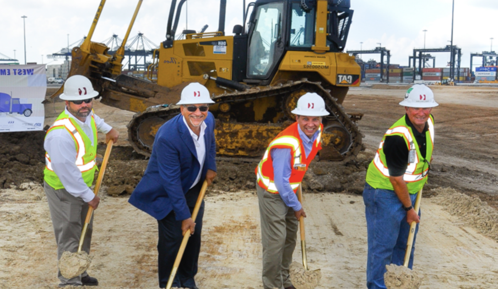 Four men in hardhats use ceremonial shovels to dig into a construction site with a bulldozer in the background