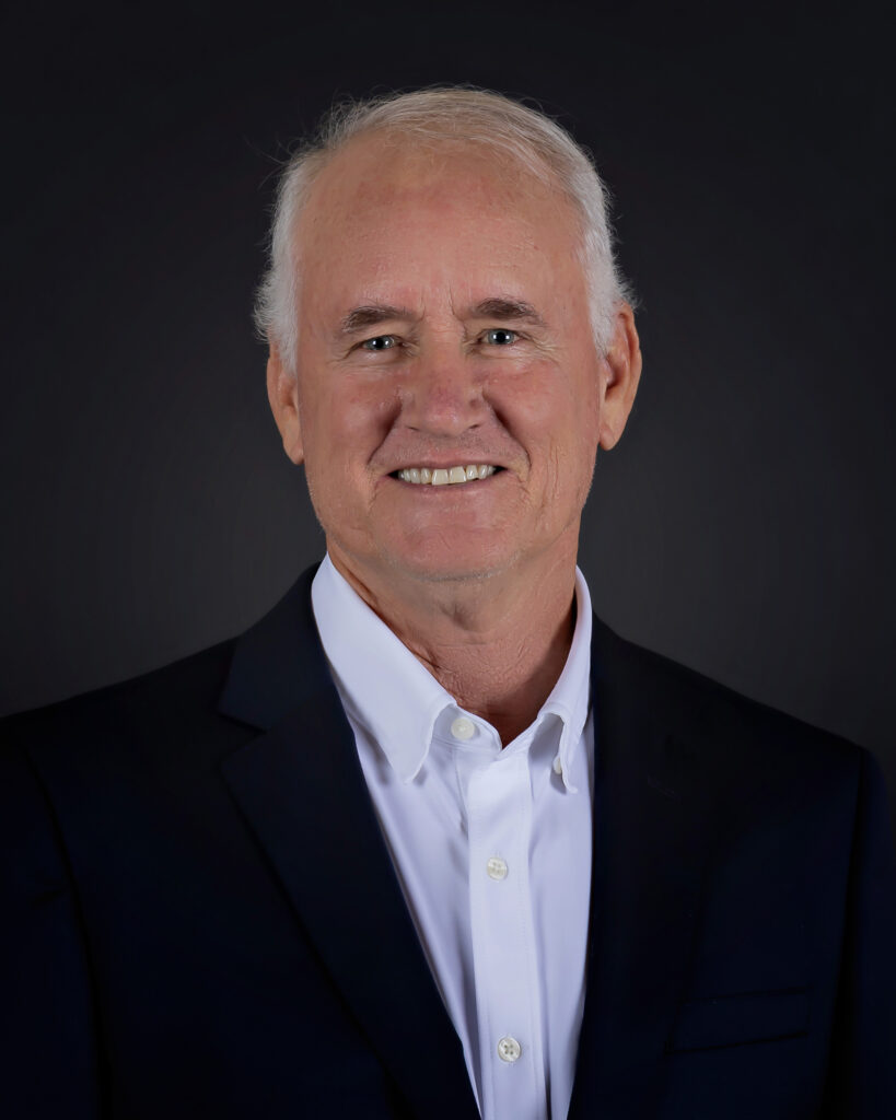 Matthew E. Kenyon, an older man with short white hair, wears a dark suit jacket and white collared shirt as he smiles against a plain dark background.
