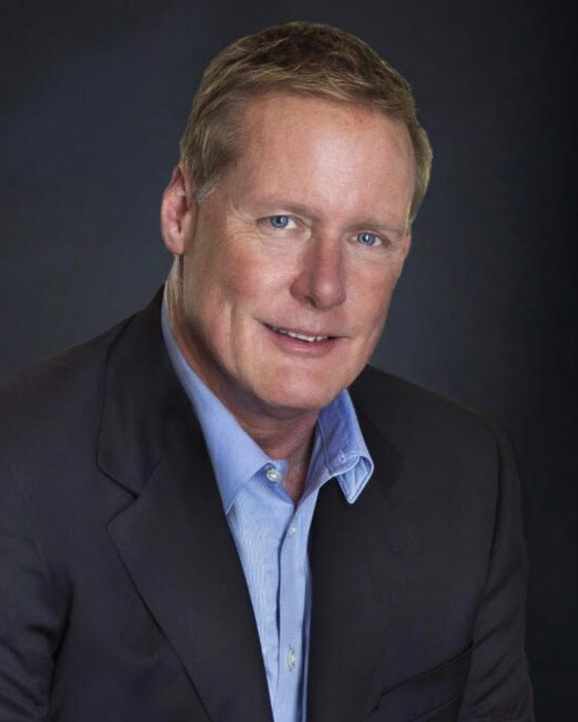 A man in a dark suit and light blue shirt is smiling in front of a dark background.