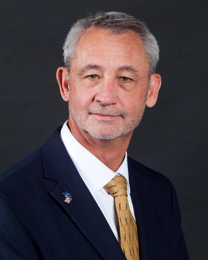 A man with gray hair and a beard wearing a navy suit, white shirt, and a gold tie, against a dark background.