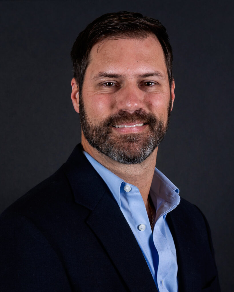A man with short dark hair and a beard, wearing a navy suit jacket and light blue shirt, poses against a dark background.