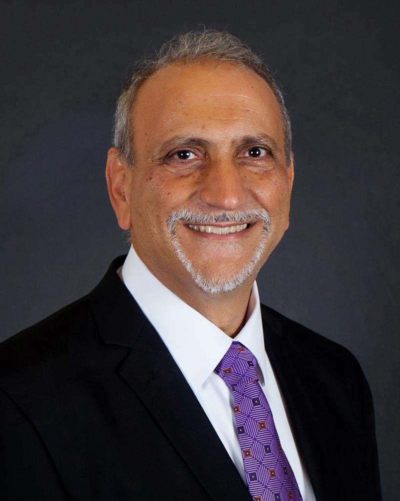 Joe Bajalia, a man in a dark suit with a purple tie, smiles against a black background