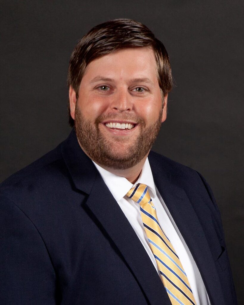 Chris Chapman, in a dark suit and yellow-striped tie, smiles against a dark background.