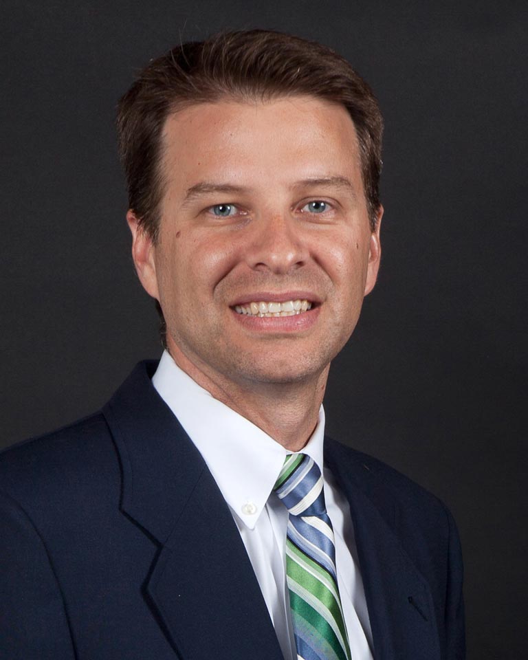 Jamison Chattin, a man with short brown hair with a green-and-blue striped tie, smiles against a dark backrgound