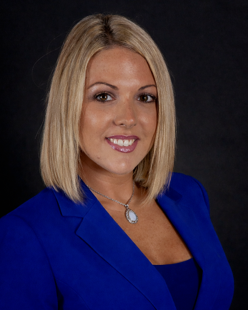 Courtney Johnson, with straight blonde hair, wears a bright blue blazer and a pendant necklace as she poses confidently against a plain dark background.