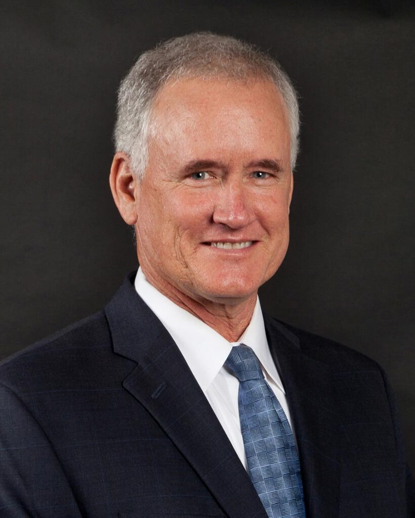 Matt Kenyon, in a suit with a light blue tie, smiles against a plain dark background.