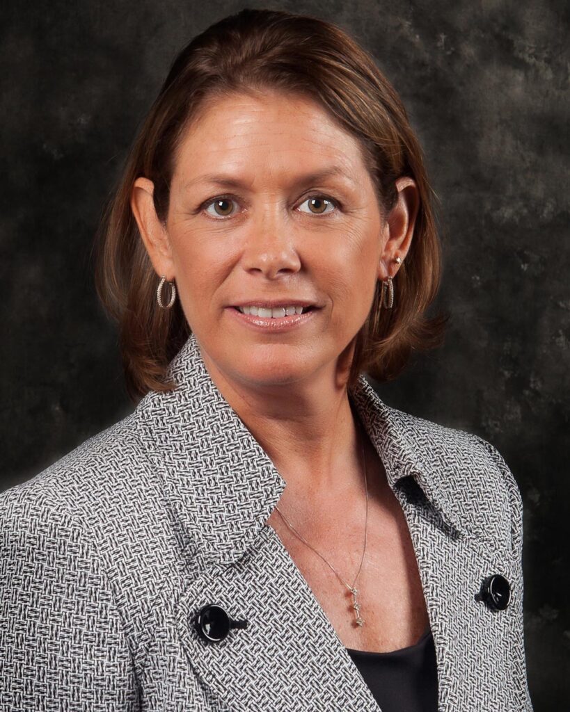 Janet Masterson, with shoulder-length hair, stands confidently against a dark background in a stylish patterned blazer.