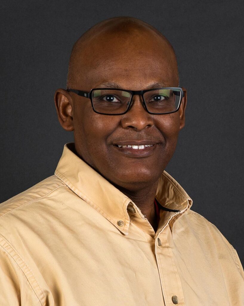 Terry Bernard, wearing glasses and a beige button-up shirt, smiles warmly against a dark background.