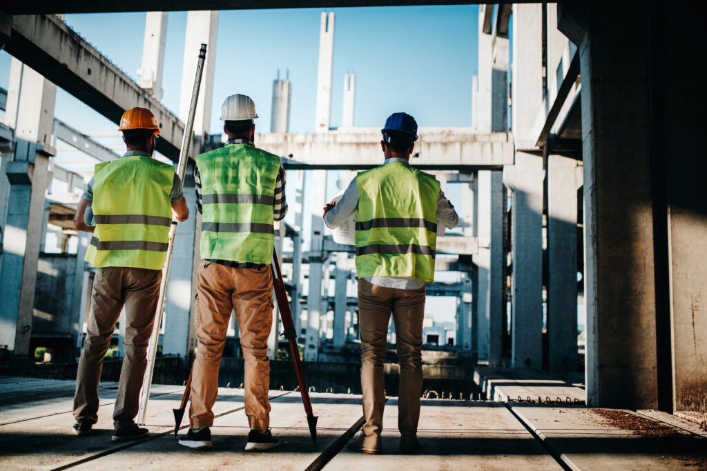 Three construction workers in reflective vests and helmets stand at a construction site, surveying the concrete framework of an unfinished structure.