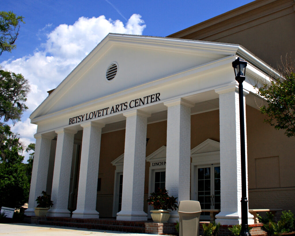 The white-columned facade of the Betsy Lovett Arts Center stands gracefully under a clear, sunny sky, framed by lush trees in the background.