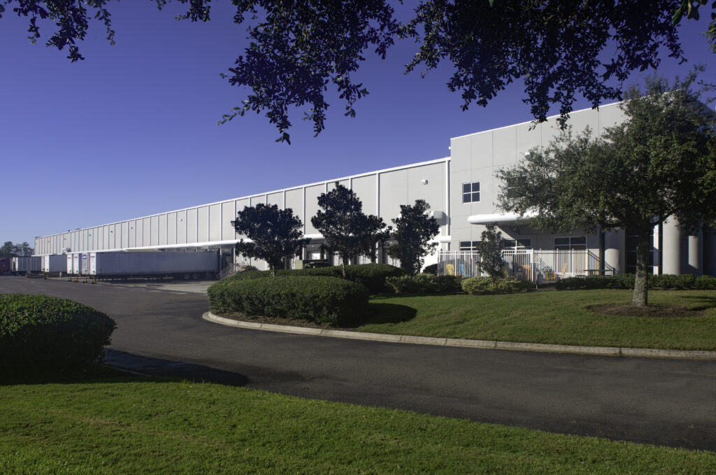 Large white warehouse building by Pattillo with loading docks, surrounded by manicured lawns, trees, and a paved driveway under a clear blue sky—ideal for future expansion.