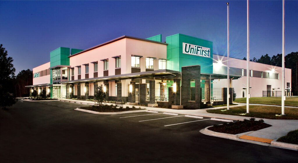 Modern UniFirst Distribution Center building at dusk with illuminated signage, large windows, and an empty parking lot in the foreground.