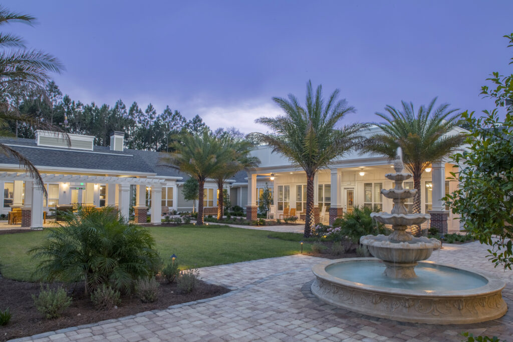 A large, single-story house with a central courtyard, palm trees, a stone fountain, and lit windows at dusk—an inviting setting reminiscent of assisted living communities like Arbor Terrace San Jose.