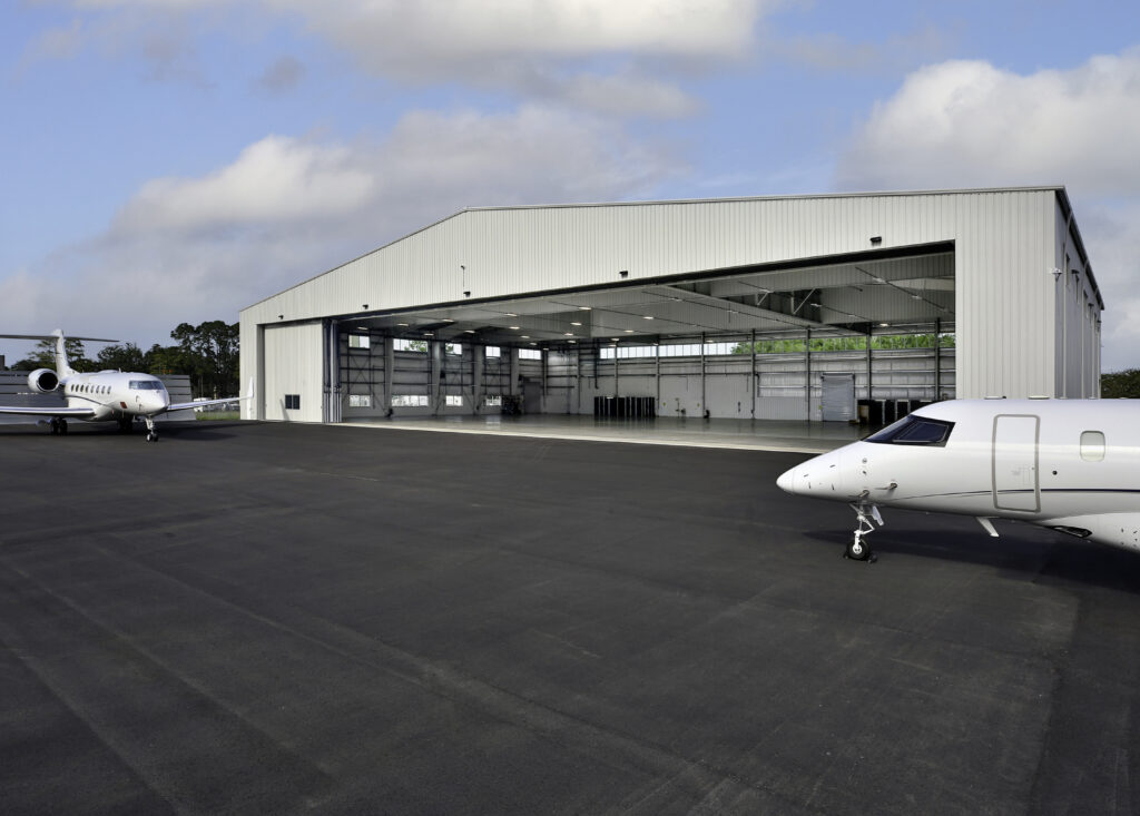 Two private jets are parked on an asphalt surface outside a large, open Signature Aviation new hangar at Jacksonville Airport under a partly cloudy sky.