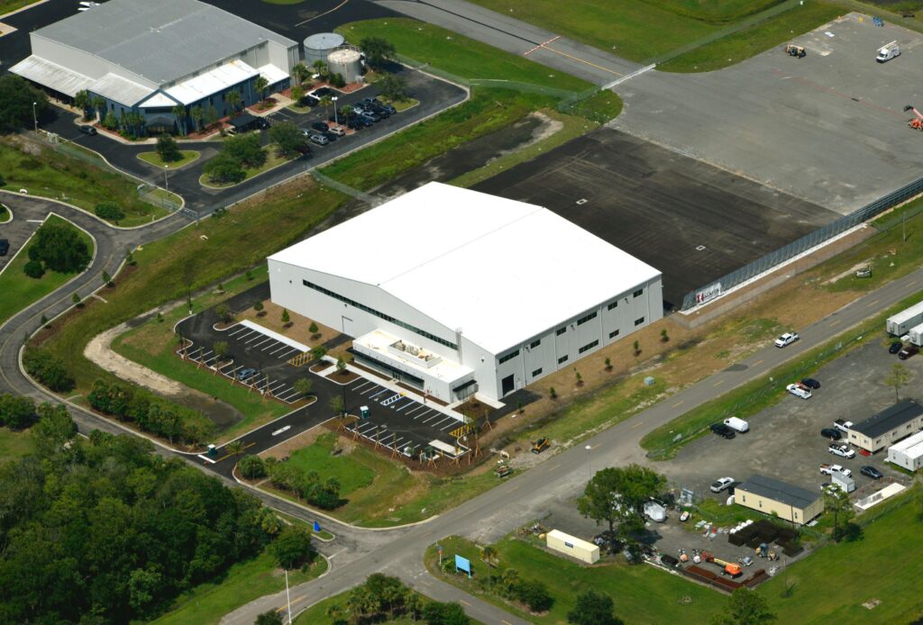 Aerial view of a large white industrial building, identified as the New Hangar for Signature Aviation at Jacksonville Airport, with adjacent parking lot, surrounded by roads, grassy areas, and smaller structures.