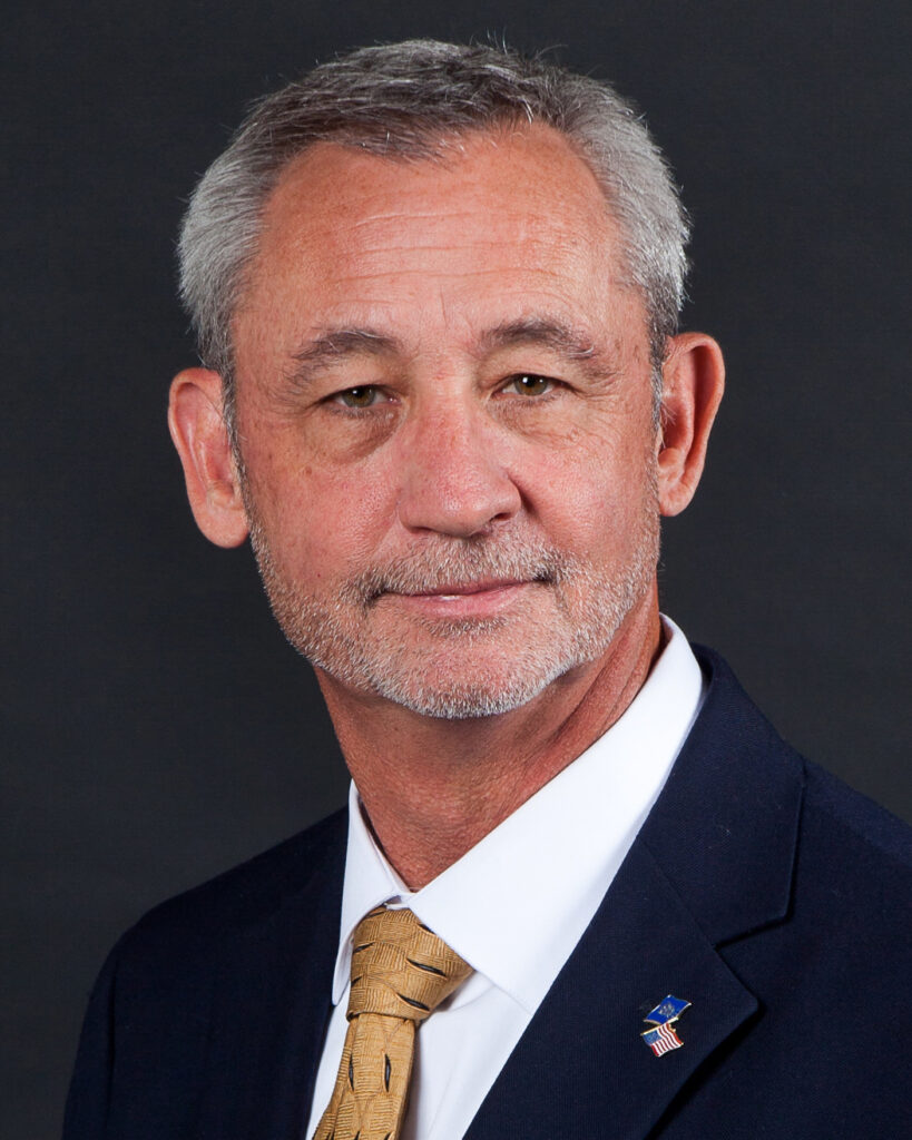 Randy Tucker, a middle-aged man with gray hair and a trimmed beard, wears a dark suit, white shirt, and yellow tie as he poses against a plain dark background.