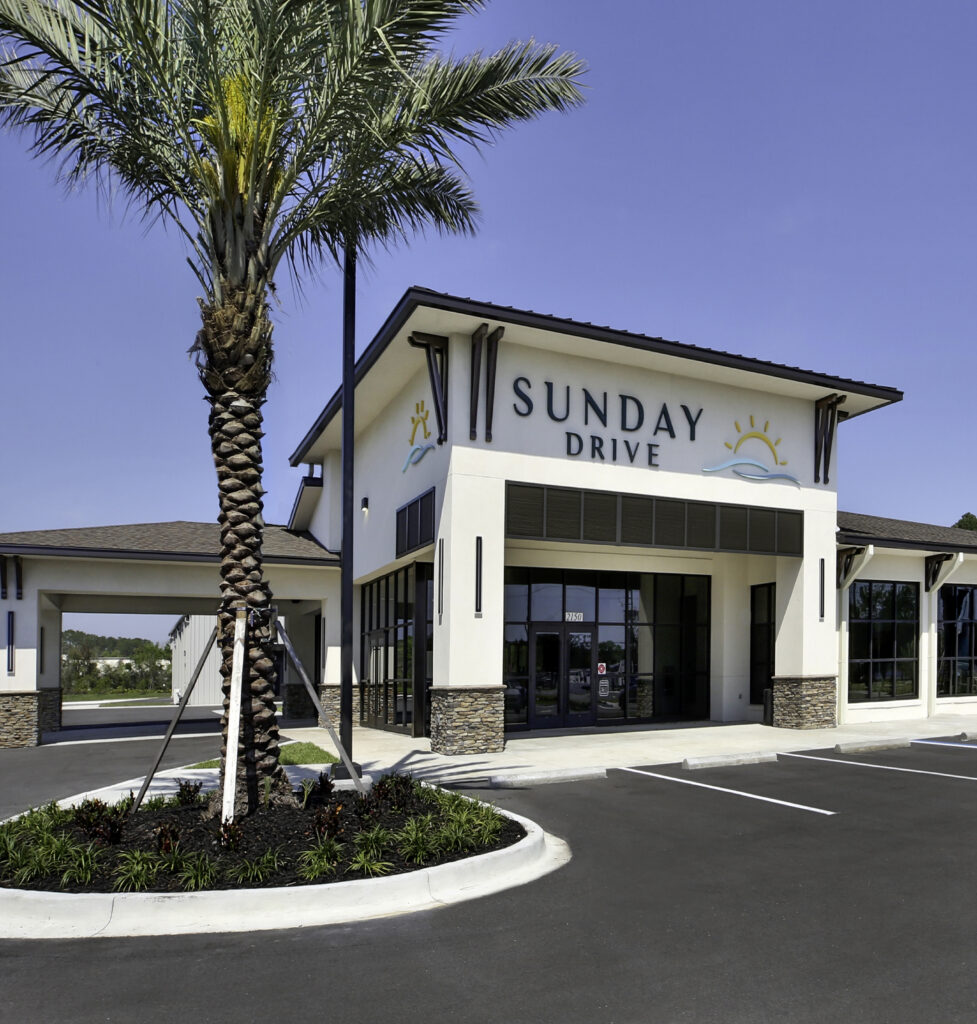 A modern commercial building labeled "Sunday Drive" stands beneath a clear sky, its large windows reflecting sunlight. A palm tree graces the front, and an empty parking lot suggests a newly opened New Car Dealership awaiting visitors.